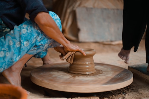 A person is shaping pottery on a spinning wheel using their hands. They are wearing vibrant blue floral-patterned pants and the setting appears earthy and artisanal.