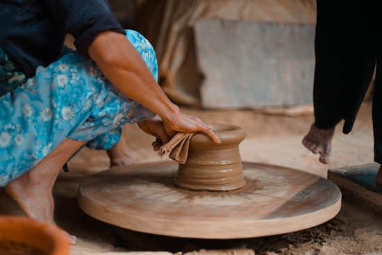 A person is shaping pottery on a spinning wheel using their hands. They are wearing vibrant blue floral-patterned pants and the setting appears earthy and artisanal.