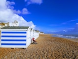 A row of beach huts with blue and white stripes is situated on a pebble beach. A stroller is parked in front of one of the huts. In the background, there's a view of the seaside town with buildings and a clear, blue sky. The sea is on the right with waves gently hitting the shore.