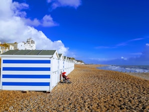 A row of beach huts with blue and white stripes is situated on a pebble beach. A stroller is parked in front of one of the huts. In the background, there's a view of the seaside town with buildings and a clear, blue sky. The sea is on the right with waves gently hitting the shore.
