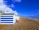 A row of beach huts with blue and white stripes is situated on a pebble beach. A stroller is parked in front of one of the huts. In the background, there's a view of the seaside town with buildings and a clear, blue sky. The sea is on the right with waves gently hitting the shore.