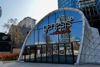 A modern building facade featuring the name 'Genesis' in stylized lettering above large glass windows. The architecture includes a geometric white frame and reflections of trees and other structures in the windows. Nearby buildings and a tree are visible in the background.