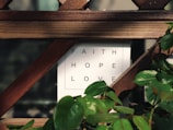 A wooden trellis partially covers a white sign with the words 'FAITH', 'HOPE', and 'LOVE' in black capital letters. Green leaves from a nearby plant frame the edges of the sign, adding a touch of nature to the scene.