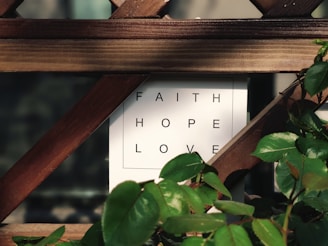 A wooden trellis partially covers a white sign with the words 'FAITH', 'HOPE', and 'LOVE' in black capital letters. Green leaves from a nearby plant frame the edges of the sign, adding a touch of nature to the scene.