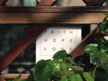 A wooden trellis partially covers a white sign with the words 'FAITH', 'HOPE', and 'LOVE' in black capital letters. Green leaves from a nearby plant frame the edges of the sign, adding a touch of nature to the scene.
