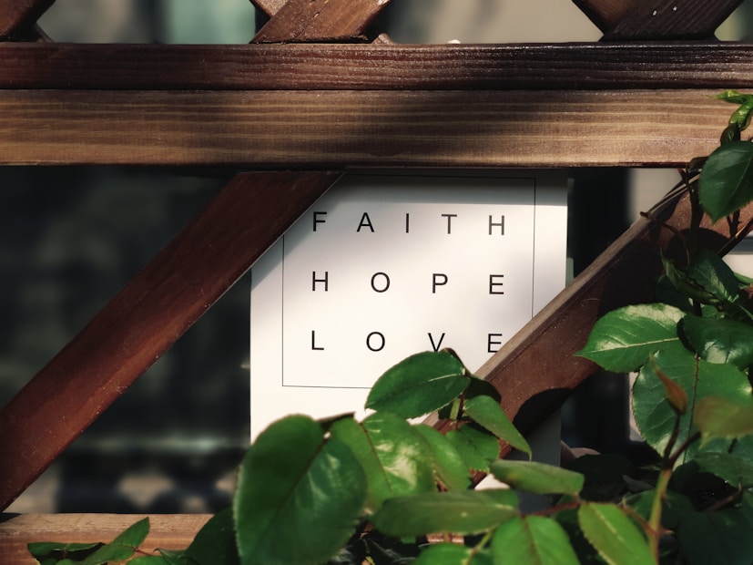 A wooden trellis partially covers a white sign with the words 'FAITH', 'HOPE', and 'LOVE' in black capital letters. Green leaves from a nearby plant frame the edges of the sign, adding a touch of nature to the scene.