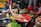 A bustling market scene with a woman crouched on the ground, reaching into a basket full of raw chickens. Fresh produce surrounds her, including bananas, leafy greens, and other vegetables. Brightly colored clothing and various containers add to the vibrant atmosphere.