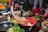 A bustling market scene with a woman crouched on the ground, reaching into a basket full of raw chickens. Fresh produce surrounds her, including bananas, leafy greens, and other vegetables. Brightly colored clothing and various containers add to the vibrant atmosphere.