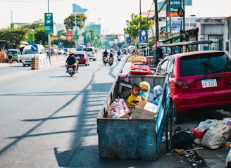 A young child sits in a cart full of discarded materials and boxes on a busy urban street. Several vehicles, including motorcycles and cars, move along the road lined with commercial buildings and advertisements. The cart is parked beside a red car, and trash is scattered on the ground nearby. The scene captures the contrast between the bustling street and the child's surrounding environment.