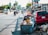 A young child sits in a cart full of discarded materials and boxes on a busy urban street. Several vehicles, including motorcycles and cars, move along the road lined with commercial buildings and advertisements. The cart is parked beside a red car, and trash is scattered on the ground nearby. The scene captures the contrast between the bustling street and the child's surrounding environment.
