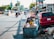 A young child sits in a cart full of discarded materials and boxes on a busy urban street. Several vehicles, including motorcycles and cars, move along the road lined with commercial buildings and advertisements. The cart is parked beside a red car, and trash is scattered on the ground nearby. The scene captures the contrast between the bustling street and the child's surrounding environment.