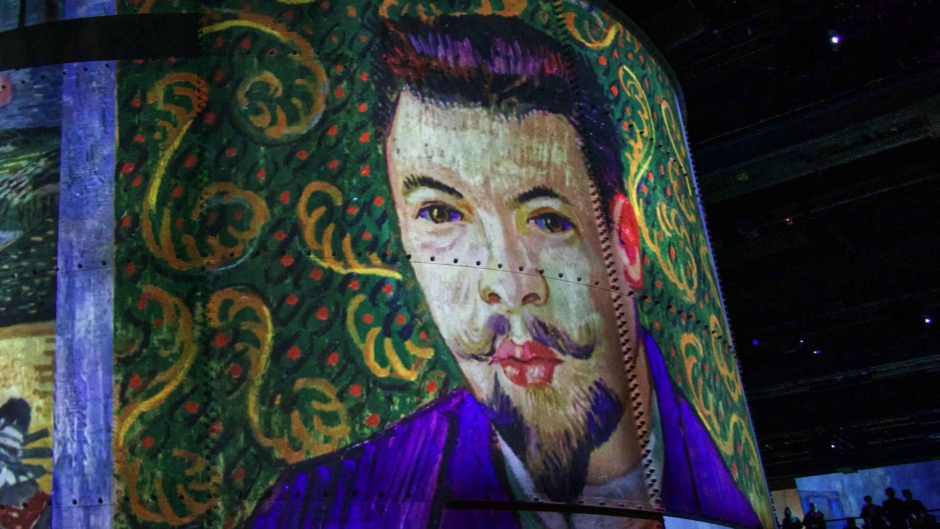 A large, vividly colored painting of a man with a distinctive mustache and beard is displayed on a cylindrical structure. The background features intricate floral patterns in green and orange, giving a sense of depth and texture.