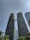 Two tall, modern skyscrapers are connected by a bridge near their tops. The buildings are covered with greenery, resembling a vertical garden. At the base, lush green trees and plants integrate with the architecture. The sky is clear with a few clouds, adding to the modern and eco-friendly feel of the scene.