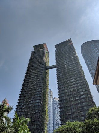 Two tall, modern skyscrapers are connected by a bridge near their tops. The buildings are covered with greenery, resembling a vertical garden. At the base, lush green trees and plants integrate with the architecture. The sky is clear with a few clouds, adding to the modern and eco-friendly feel of the scene.
