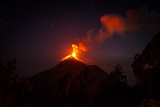 A volcano is erupting at night, with bright orange lava glowing at the summit and dark smoke billowing into the starry sky. The surrounding landscape is silhouetted against the fiery glow with trees visible in the foreground.