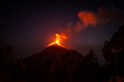 A volcano is erupting at night, with bright orange lava glowing at the summit and dark smoke billowing into the starry sky. The surrounding landscape is silhouetted against the fiery glow with trees visible in the foreground.