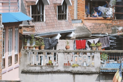 A residential scene with brick and plaster buildings featuring rooftop terraces and architectural details. Several potted plants are placed on a concrete railing. Clothes are hung out to dry on a line strung between structures. Roofs are covered with tiles and corrugated metal sheets.