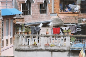 A residential scene with brick and plaster buildings featuring rooftop terraces and architectural details. Several potted plants are placed on a concrete railing. Clothes are hung out to dry on a line strung between structures. Roofs are covered with tiles and corrugated metal sheets.