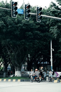 A bustling city street intersection with a traffic light displaying red signals. Several people are gathered at the crosswalk, including a person on a motorcycle wearing an orange helmet, and others standing or walking with bicycles. Lush green trees form a backdrop, and a car is partially visible behind the trees. Traffic signs and a security camera are mounted on poles.