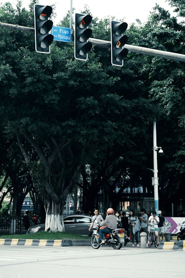 A bustling city street intersection with a traffic light displaying red signals. Several people are gathered at the crosswalk, including a person on a motorcycle wearing an orange helmet, and others standing or walking with bicycles. Lush green trees form a backdrop, and a car is partially visible behind the trees. Traffic signs and a security camera are mounted on poles.