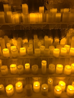 Numerous lit candles of varying heights are organized on several shelves, with wax dripping down the sides, creating an ambient and warm glow.