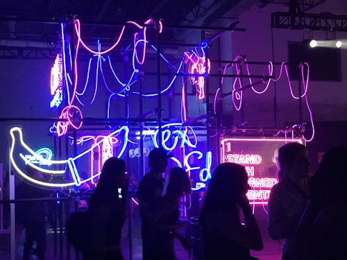 A series of vibrant neon lights in purple, pink, and blue are displayed, forming abstract shapes and letters. Silhouettes of people are visible in the foreground, appearing to observe the colorful installation. The environment is dimly lit, enhancing the intensity of the neon glow.