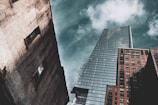 A perspective shot of three skyscrapers against a partly cloudy sky. The tallest building in the center has a modern glass facade with a sharp architectural design. On the right, a brick building displays a 'Rent Now' sign with a phone number. On the left, a weathered concrete building features two visible window openings.