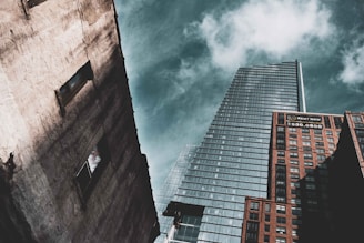 A perspective shot of three skyscrapers against a partly cloudy sky. The tallest building in the center has a modern glass facade with a sharp architectural design. On the right, a brick building displays a 'Rent Now' sign with a phone number. On the left, a weathered concrete building features two visible window openings.