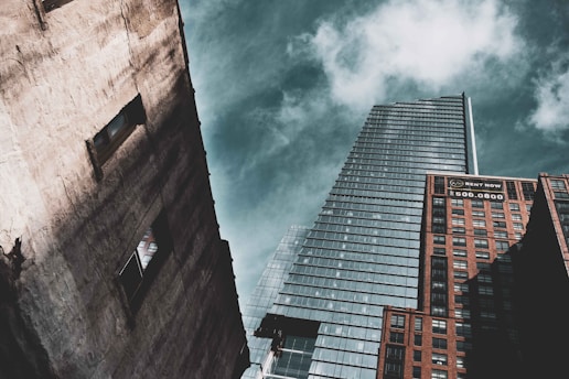 A perspective shot of three skyscrapers against a partly cloudy sky. The tallest building in the center has a modern glass facade with a sharp architectural design. On the right, a brick building displays a 'Rent Now' sign with a phone number. On the left, a weathered concrete building features two visible window openings.
