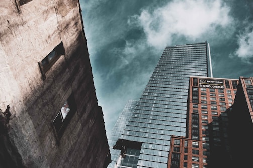 A perspective shot of three skyscrapers against a partly cloudy sky. The tallest building in the center has a modern glass facade with a sharp architectural design. On the right, a brick building displays a 'Rent Now' sign with a phone number. On the left, a weathered concrete building features two visible window openings.