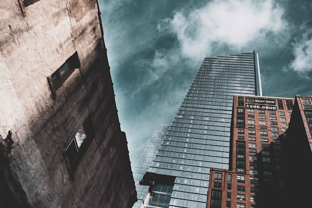 A perspective shot of three skyscrapers against a partly cloudy sky. The tallest building in the center has a modern glass facade with a sharp architectural design. On the right, a brick building displays a 'Rent Now' sign with a phone number. On the left, a weathered concrete building features two visible window openings.