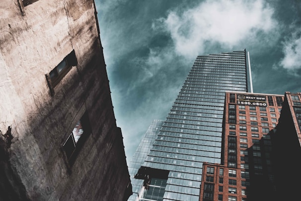 A perspective shot of three skyscrapers against a partly cloudy sky. The tallest building in the center has a modern glass facade with a sharp architectural design. On the right, a brick building displays a 'Rent Now' sign with a phone number. On the left, a weathered concrete building features two visible window openings.