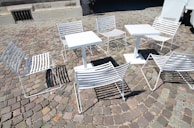 White metal chairs and tables are arranged on a cobblestone pavement, casting shadows on the ground. The setting suggests an outdoor café or public seating area, with a mix of sunlight and shade affecting the visibility of details.