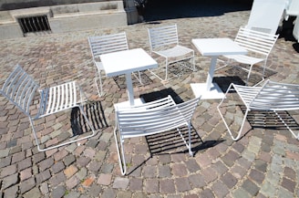 White metal chairs and tables are arranged on a cobblestone pavement, casting shadows on the ground. The setting suggests an outdoor café or public seating area, with a mix of sunlight and shade affecting the visibility of details.