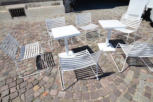 White metal chairs and tables are arranged on a cobblestone pavement, casting shadows on the ground. The setting suggests an outdoor café or public seating area, with a mix of sunlight and shade affecting the visibility of details.