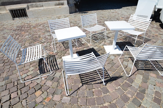 White metal chairs and tables are arranged on a cobblestone pavement, casting shadows on the ground. The setting suggests an outdoor café or public seating area, with a mix of sunlight and shade affecting the visibility of details.