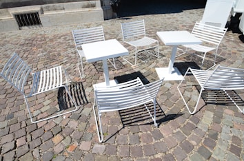 White metal chairs and tables are arranged on a cobblestone pavement, casting shadows on the ground. The setting suggests an outdoor café or public seating area, with a mix of sunlight and shade affecting the visibility of details.