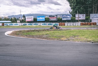 A go-kart with a racing driver in a helmet is taking a turn on an outdoor track surrounded by barriers. Advertisements and greenery are visible in the background under a cloudy sky.
