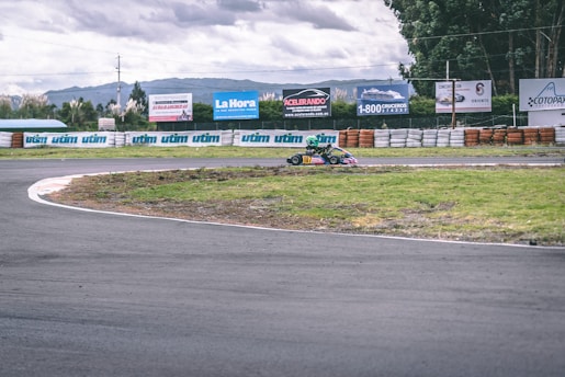 A go-kart with a racing driver in a helmet is taking a turn on an outdoor track surrounded by barriers. Advertisements and greenery are visible in the background under a cloudy sky.
