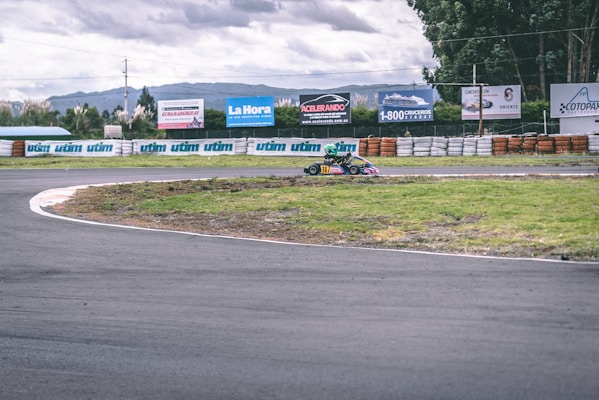 A go-kart with a racing driver in a helmet is taking a turn on an outdoor track surrounded by barriers. Advertisements and greenery are visible in the background under a cloudy sky.