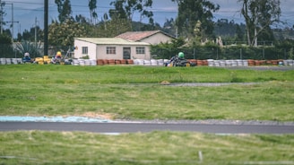 A go-kart race is taking place on a track surrounded by lush green grass and safety barriers made of tires. In the background, a house and trees are visible under an overcast sky, with racers wearing helmets navigating the course.