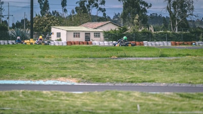 A go-kart race is taking place on a track surrounded by lush green grass and safety barriers made of tires. In the background, a house and trees are visible under an overcast sky, with racers wearing helmets navigating the course.