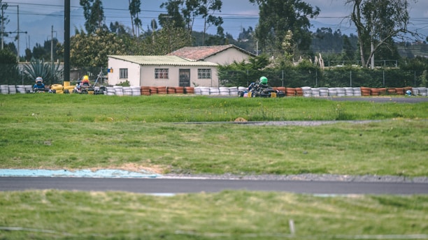 A go-kart race is taking place on a track surrounded by lush green grass and safety barriers made of tires. In the background, a house and trees are visible under an overcast sky, with racers wearing helmets navigating the course.