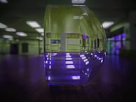 A close-up of a transparent, curved object, likely a glass or acrylic piece, is centrally placed on a reflective surface. The background features bright, rectangular ceiling lights and a room with blurred walls, giving a sense of depth. The reflection causes a light pattern to appear on the surface beneath the object, creating an interesting visual effect.