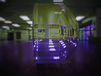 A close-up of a transparent, curved object, likely a glass or acrylic piece, is centrally placed on a reflective surface. The background features bright, rectangular ceiling lights and a room with blurred walls, giving a sense of depth. The reflection causes a light pattern to appear on the surface beneath the object, creating an interesting visual effect.