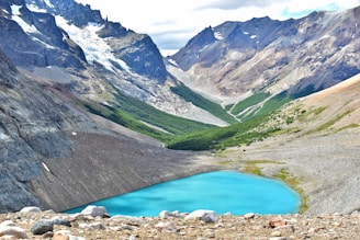 A vibrant blue mountain lake is nestled in a rocky valley surrounded by towering, rugged peaks. The mountains have patches of snow and ice, contrasting with areas of green vegetation. The sky above is partially cloudy, adding depth to the scene.