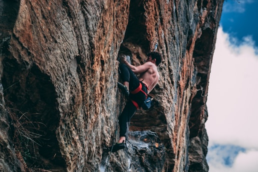 A rock climber wearing a harness and climbing shoes is ascending a steep rock face. The climber has an athletic build and focuses intently on maintaining grip and balance while scaling the rough, textured surface of the cliff. The sky in the background is partly cloudy, adding a contrasting backdrop to the scene.