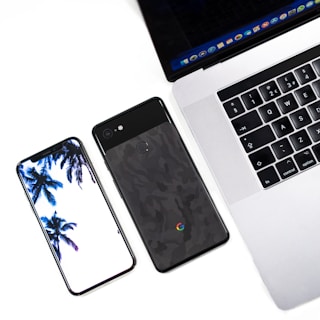 A modern workspace setup featuring a smartphone with a palm tree wallpaper, a second smartphone with a textured back, and a laptop displaying an open web browser. The devices are placed on a light surface, suggesting a clean and minimalistic style.