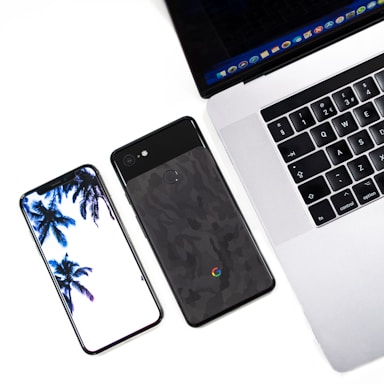 A modern workspace setup featuring a smartphone with a palm tree wallpaper, a second smartphone with a textured back, and a laptop displaying an open web browser. The devices are placed on a light surface, suggesting a clean and minimalistic style.