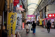 A bustling indoor shopping arcade with a variety of signs and advertisements written in Japanese. The setting features a high arched ceiling with supports and decorative elements. Shoppers are walking along the central pathway, surrounded by stores on either side offering various goods and services.